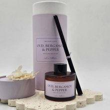 Load image into Gallery viewer, Luxury reed diffuser home fragrance in 100ml amber glass bottle with black reeds. Presented in lilac cylinder box ready for gifting. Fragrance Oud, Bergamot & Pepper