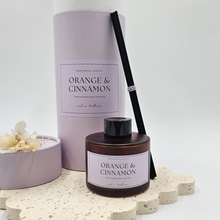 Load image into Gallery viewer, Luxury reed diffuser home fragrance in 100ml amber glass bottle with black reeds. Presented in lilac cylinder box ready for gifting. Fragrance orange and cinnamon.
