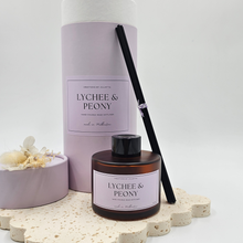 Load image into Gallery viewer, Luxury reed diffuser home fragrance in 100ml amber glass bottle with black reeds.  Presented in lilac cylinder box ready for gifting. Fragrance Lychee &amp; Peony
